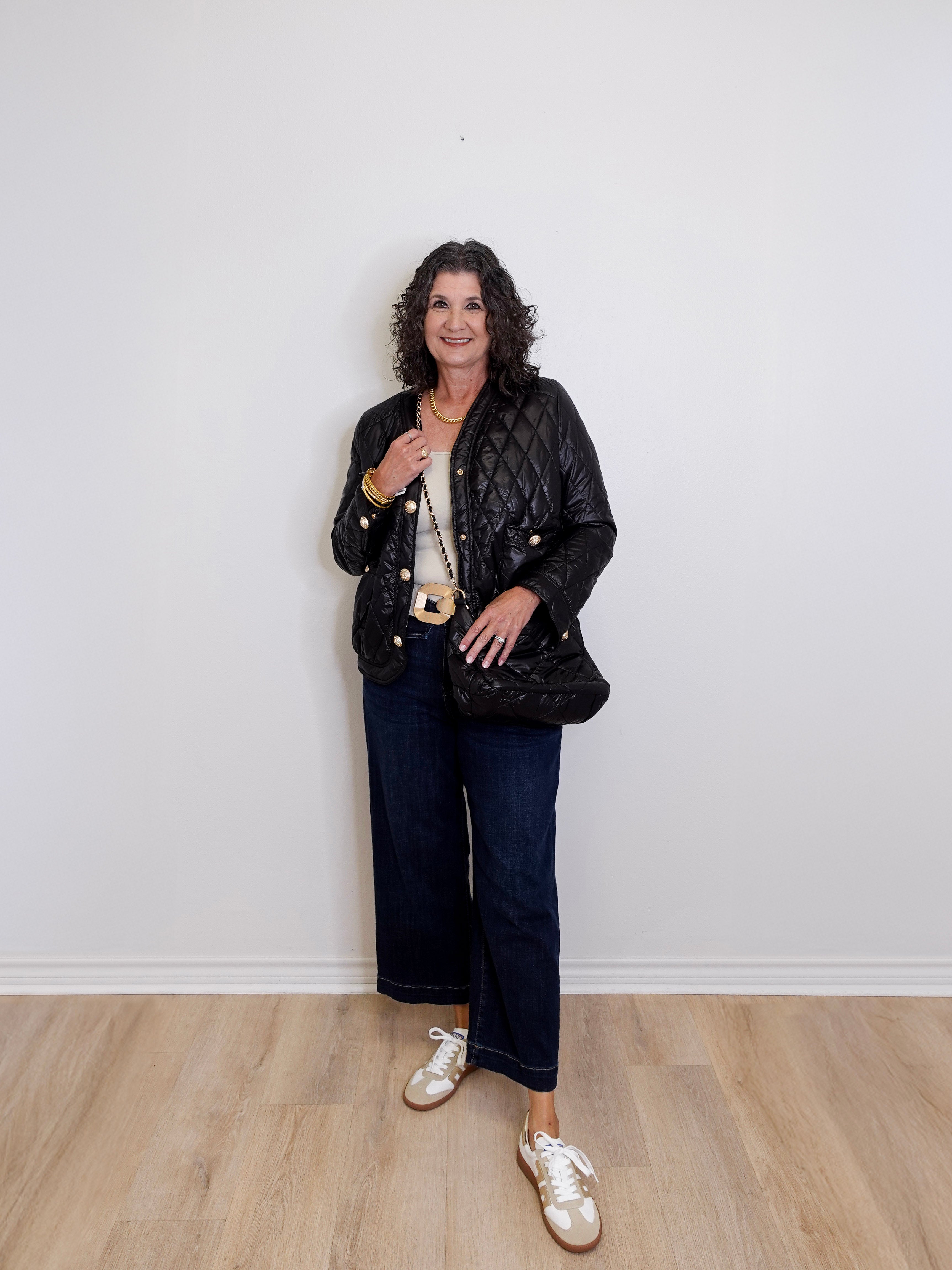 Woman wearing a black quilted jacket, white sneakers, and dark jeans against a plain white wall.