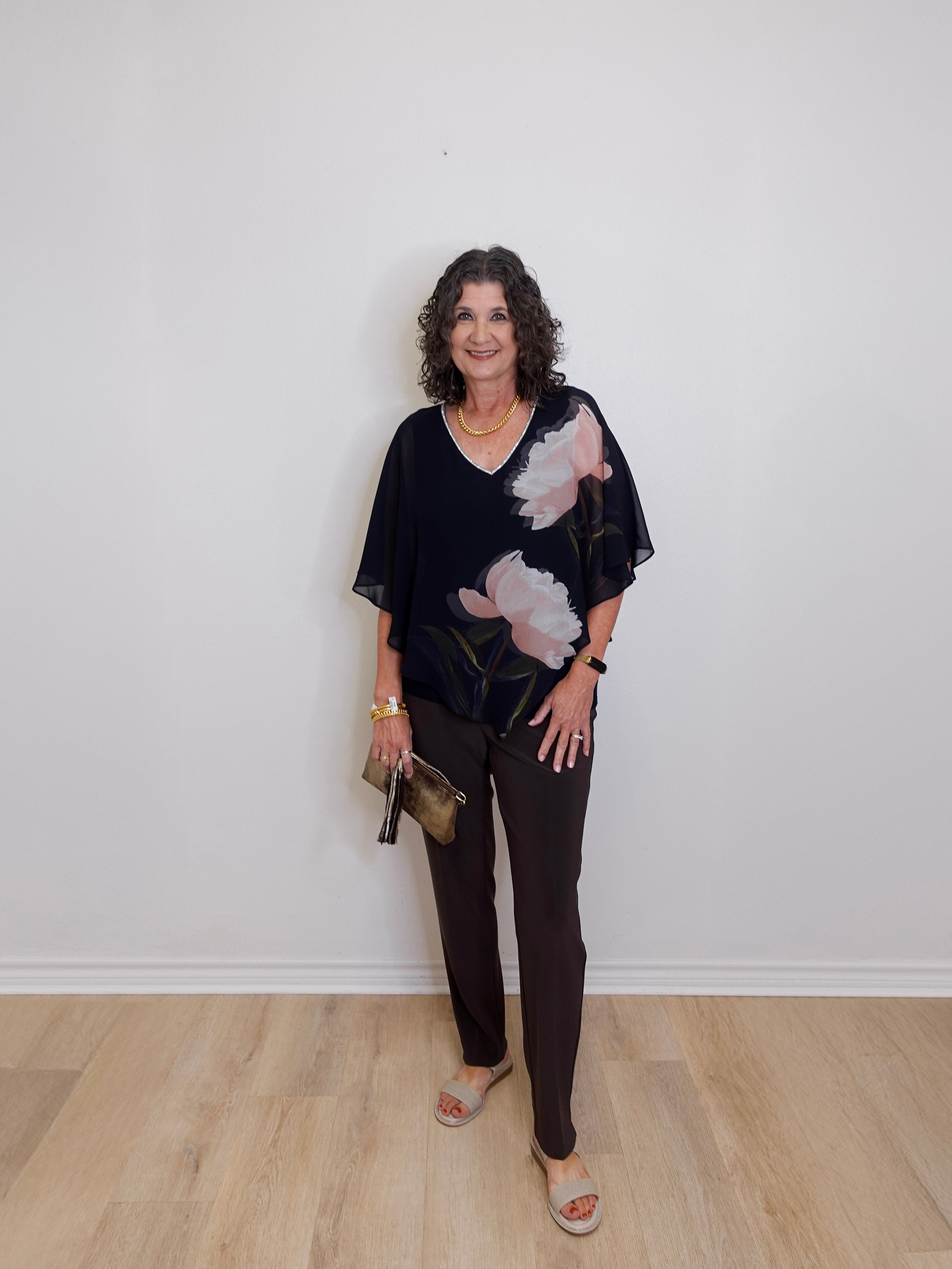 Woman wearing a black floral blouse and black pants against a white wall.
