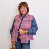 Woman wearing a patterned pink vest over a denim shirt against a white wall.