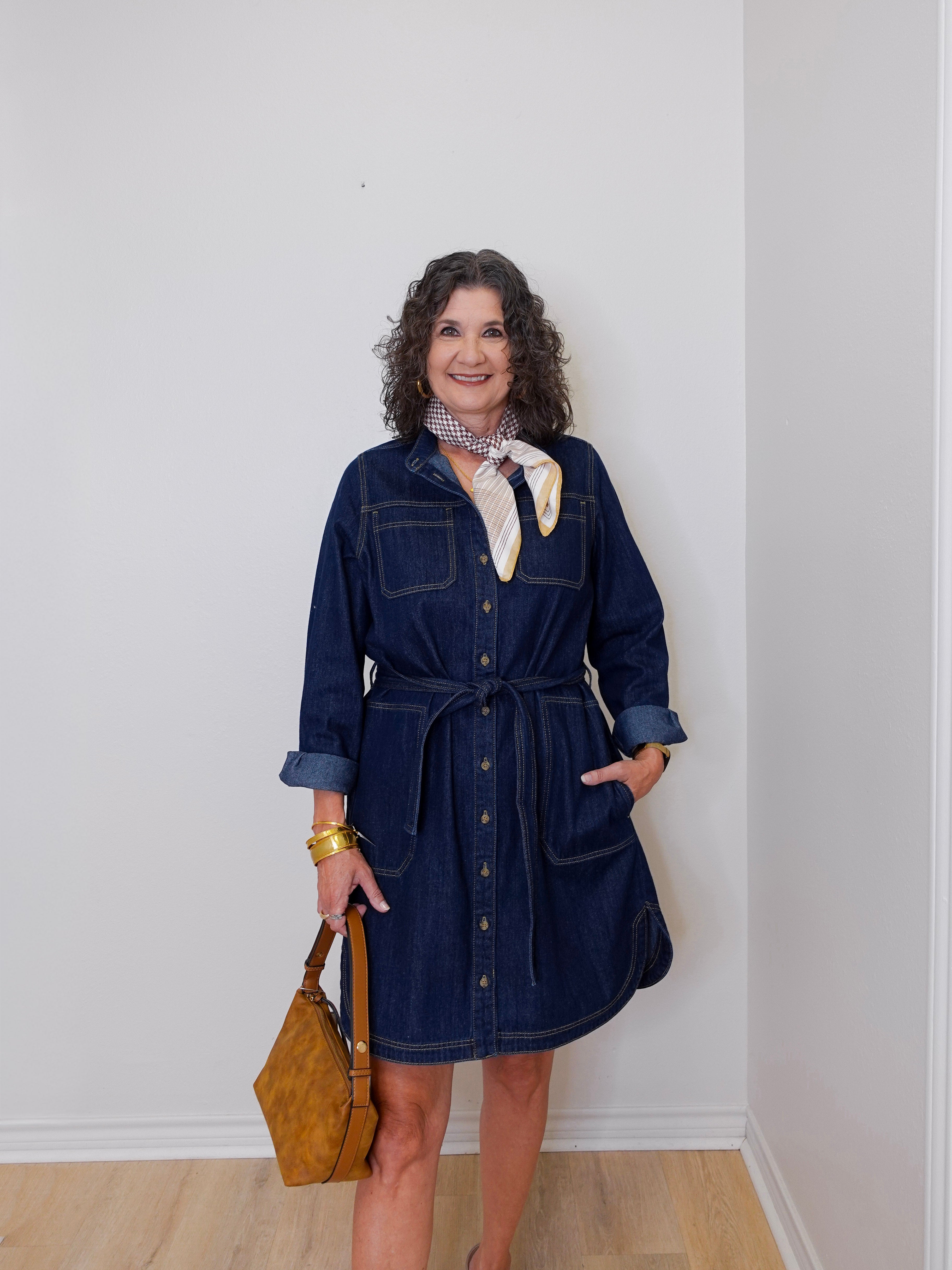 Woman wearing a denim dress with a scarf and holding a bag against a white wall.