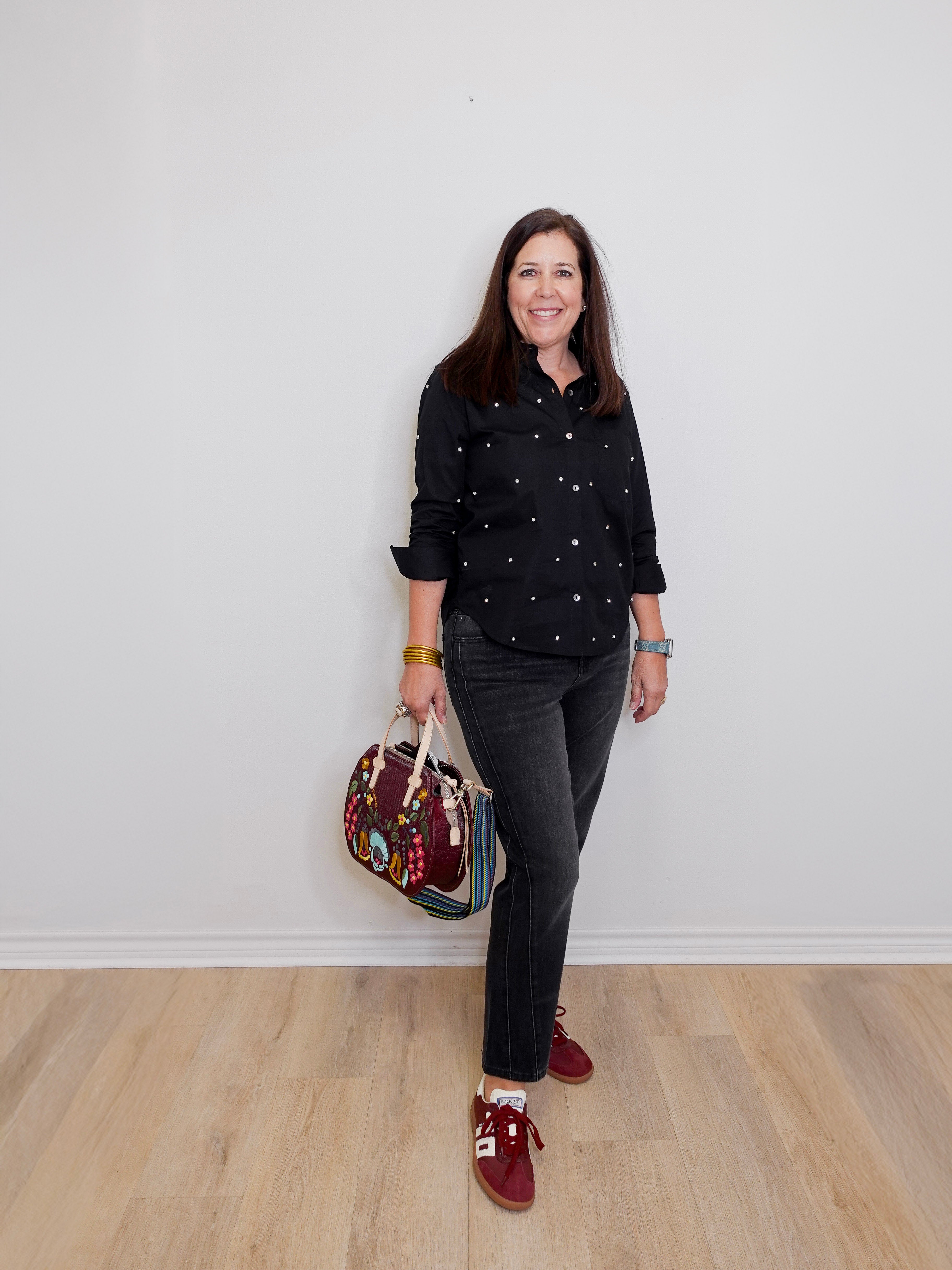Woman in black polka dot shirt and gray pants holding a colorful bag against a white wall.