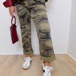 Person wearing camouflage pants and white sneakers, holding a red handbag indoors.