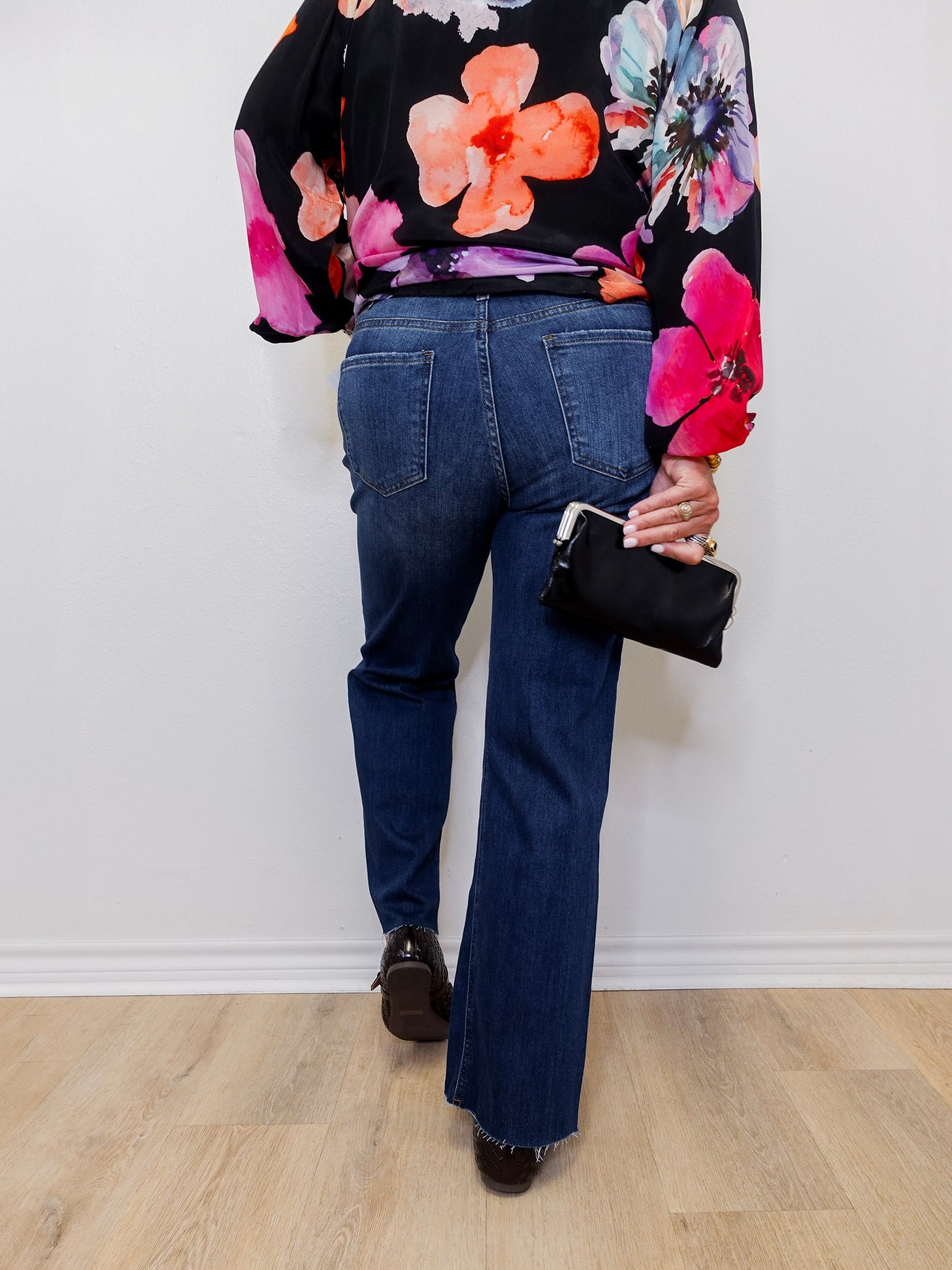 Person wearing a floral blouse and blue jeans holding a black handbag against a white wall.