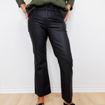 Person wearing a green shirt, black pants, and black loafers with gold accents against a white wall.