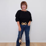 Woman wearing a black top and blue jeans with a gold belt, standing against a white wall.