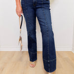 Person wearing blue jeans with a gold belt and sandals, standing on a wooden floor.