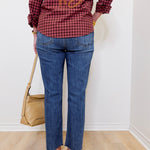 Person wearing a red plaid shirt with gold text and blue jeans, standing against a white wall.