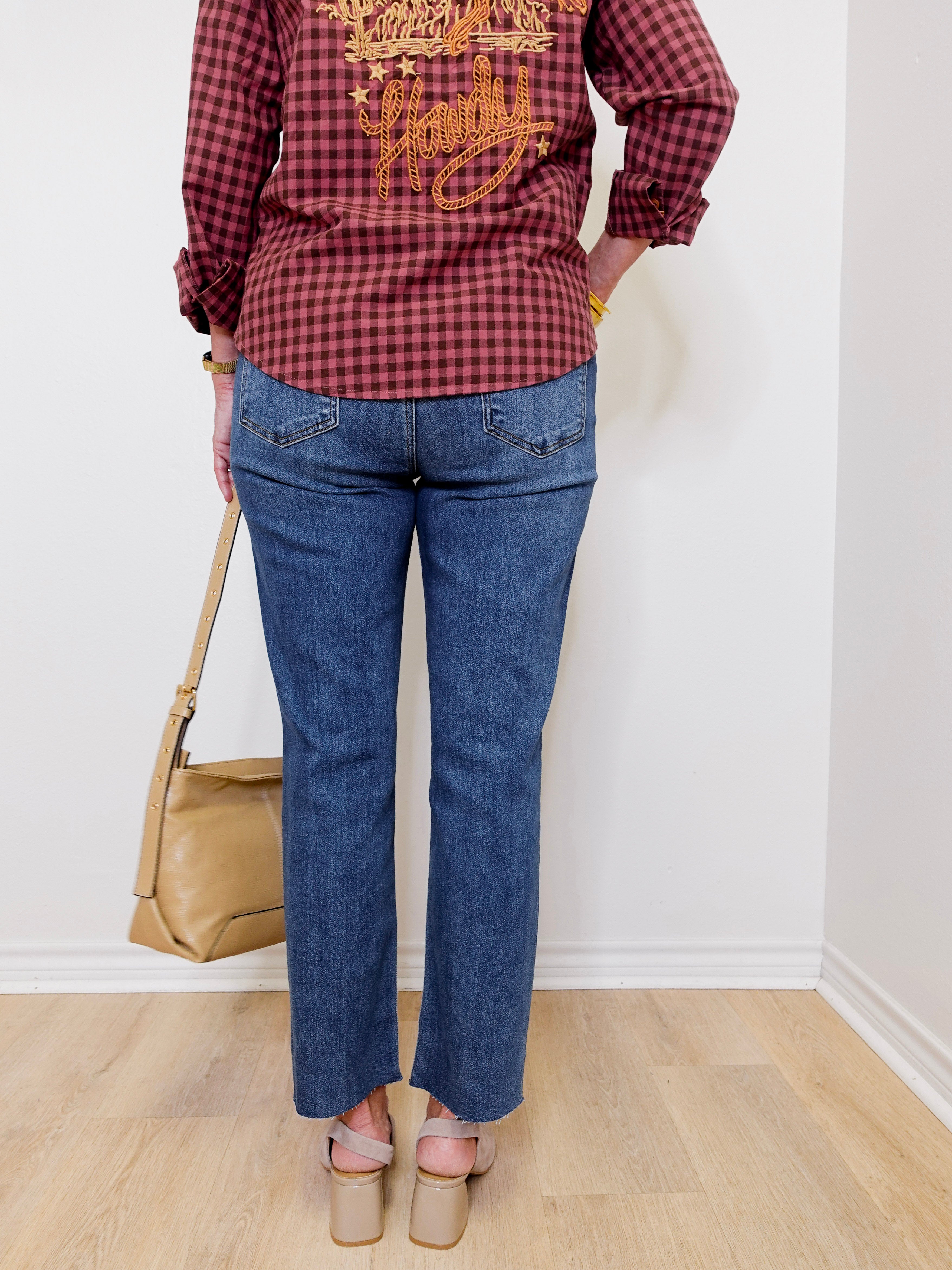 Person wearing a red plaid shirt with gold text and blue jeans, standing against a white wall.