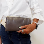 Person holding a gray clutch bag with a white shirt and dark jeans against a plain background