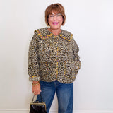 Woman wearing a leopard print jacket and blue jeans, holding a black handbag against a white wall.