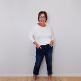 Woman wearing a white long sleeve and blue jeans standing against a plain white wall.