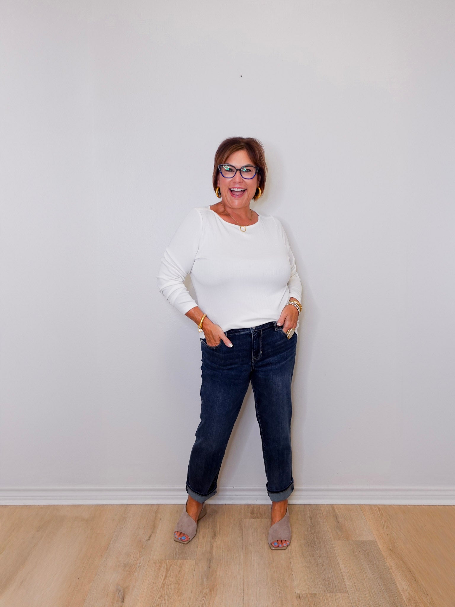 Woman wearing a white long sleeve and blue jeans standing against a plain white wall.