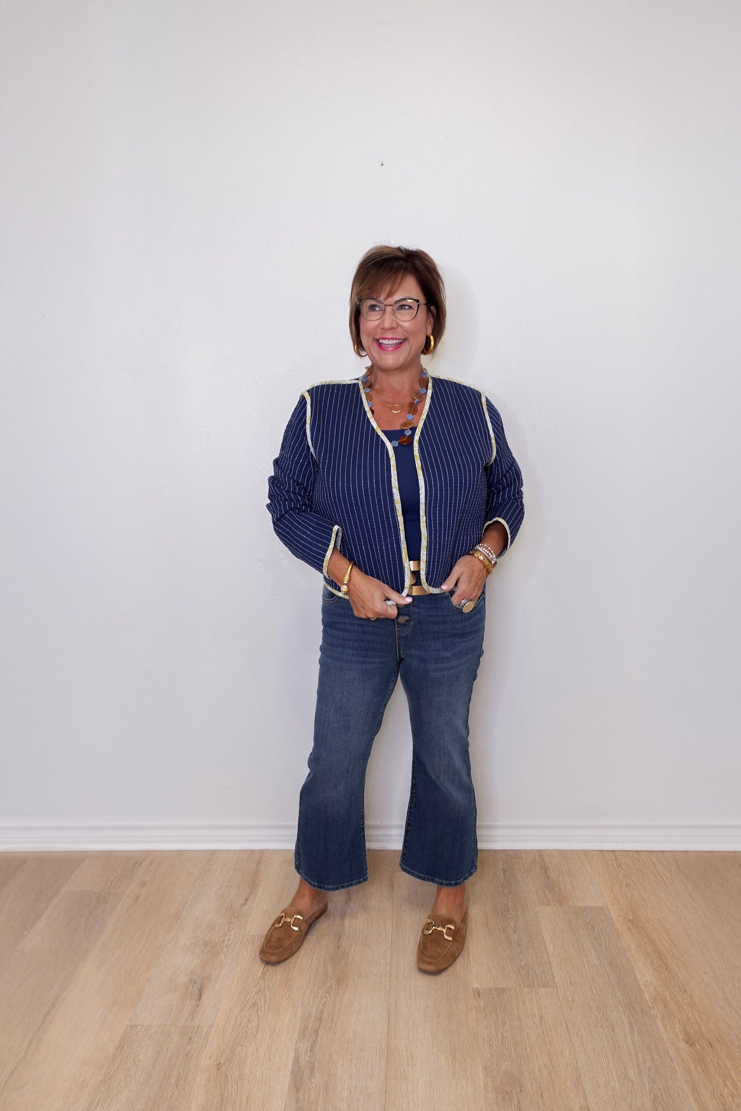 Woman wearing a blue patterned jacket and jeans standing against a white wall.
