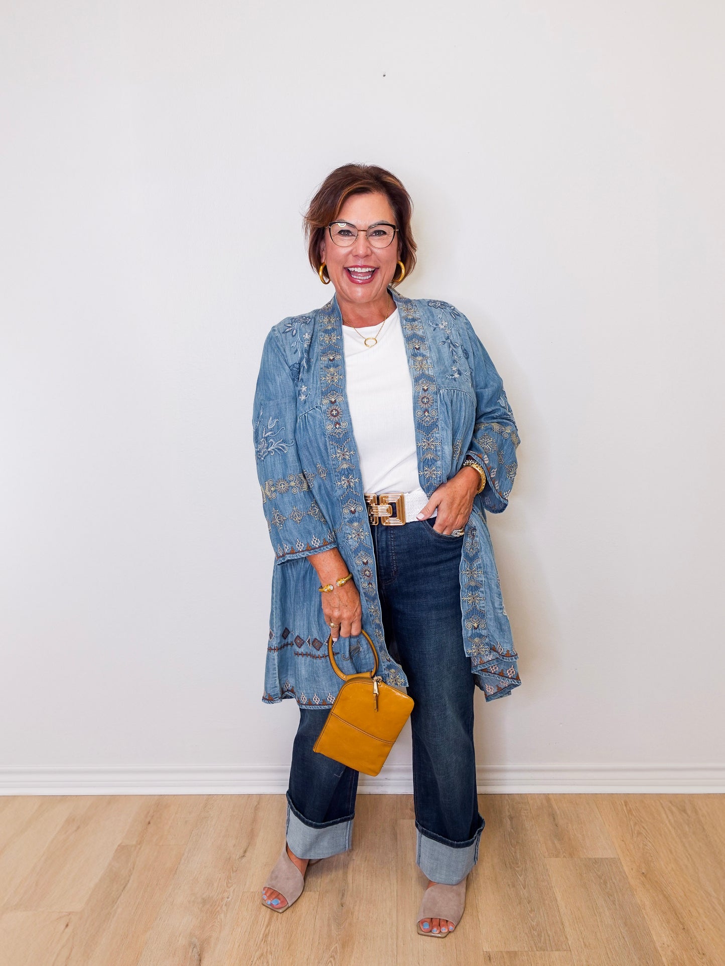 Woman wearing a denim jacket, white shirt, and jeans, holding a yellow handbag against a white wall.
