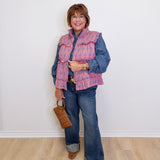 Woman wearing a pink quilted vest over a denim jacket with blue jeans and brown boots, holding a brown bag against a white background.