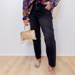 Person wearing a colorful patterned shirt, black jeans, and holding a beige handbag against a white wall.