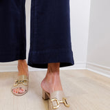 Person wearing beige sandals with gold buckles on a light wooden floor.