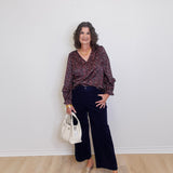 Woman wearing a patterned blouse and dark pants against a white wall.