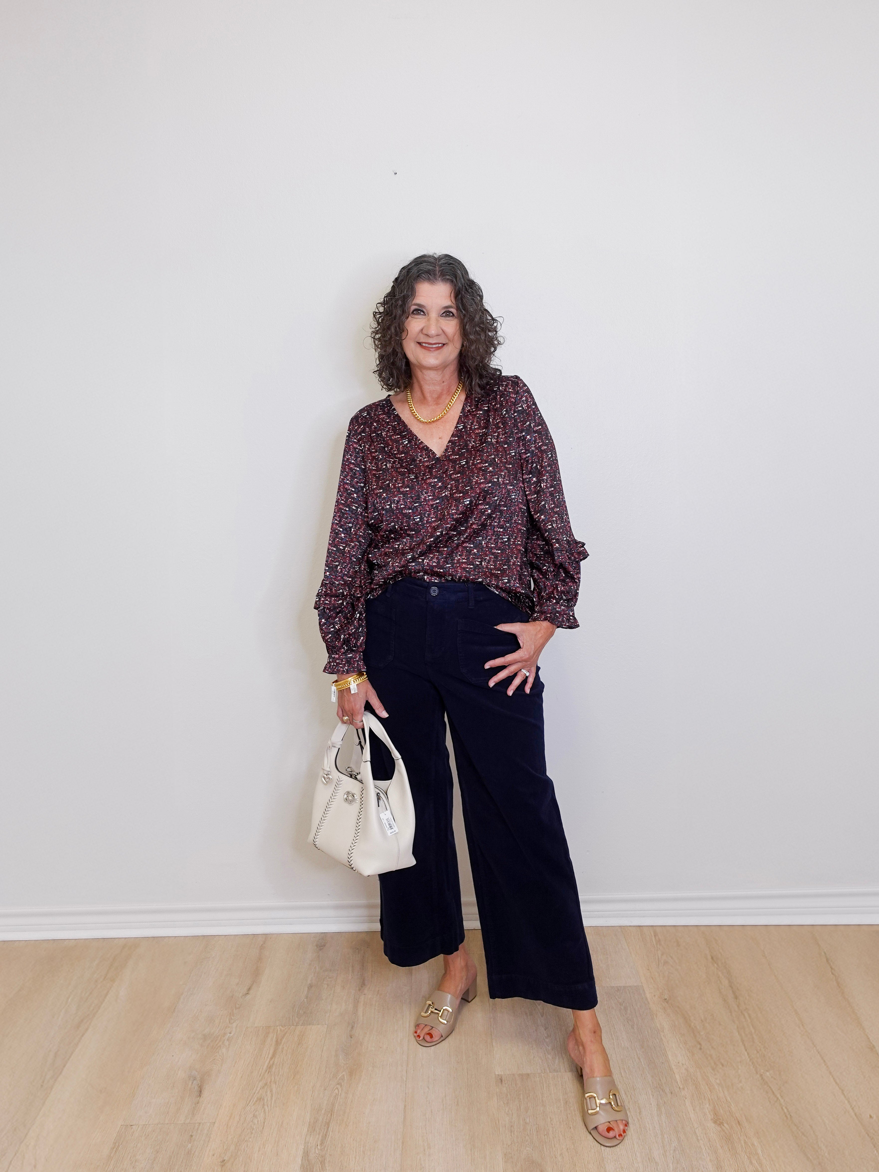 Woman wearing a patterned blouse and dark pants against a white wall.
