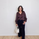 Woman standing against a white wall wearing a patterned blouse and black pants, holding a white handbag.
