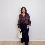 Woman standing against a white wall wearing a patterned blouse and black pants, holding a white handbag.