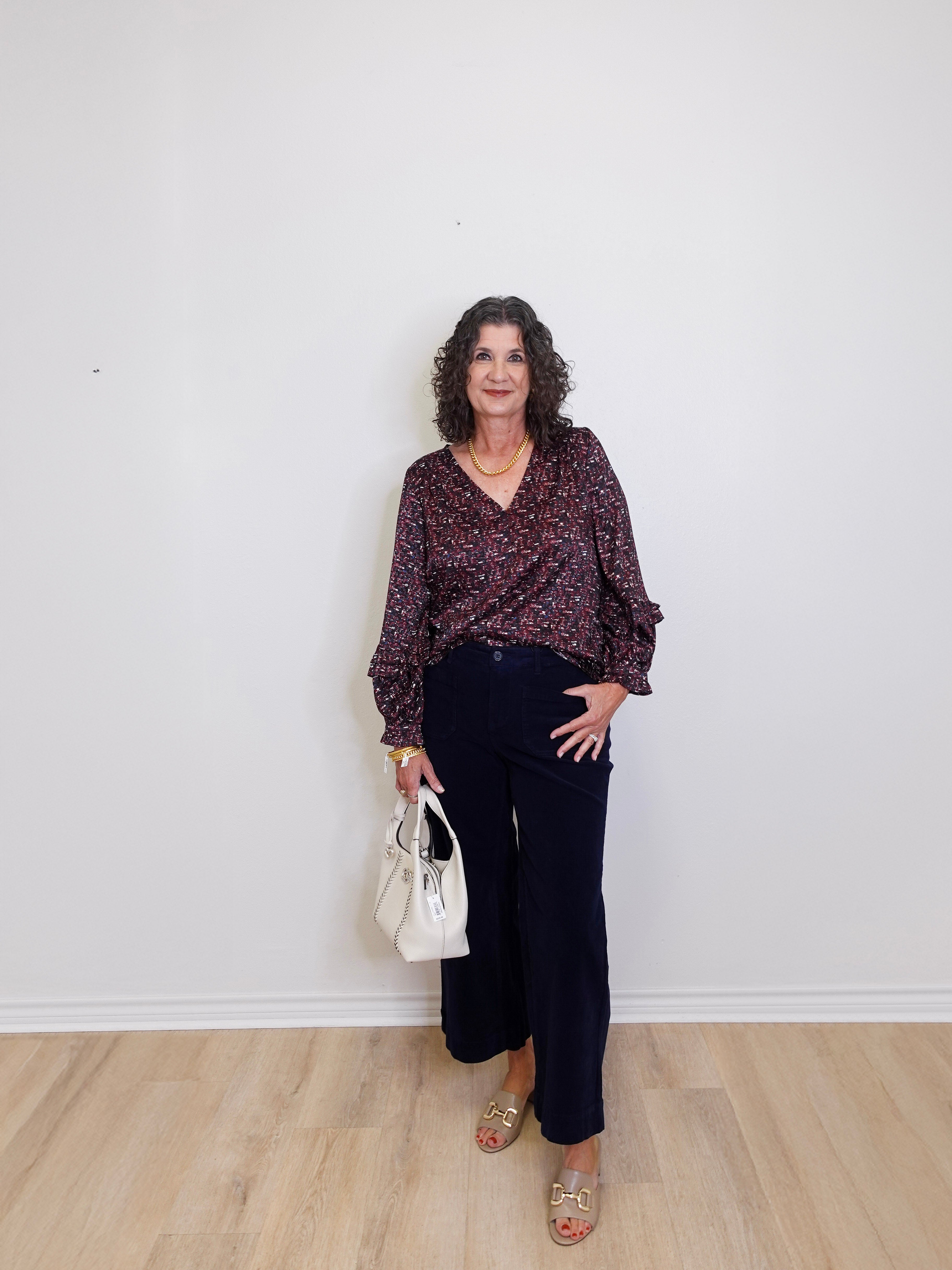 Woman standing against a white wall wearing a patterned blouse and black pants, holding a white handbag.