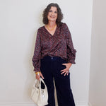 Woman wearing a patterned blouse and dark pants, holding a white handbag against a plain background
