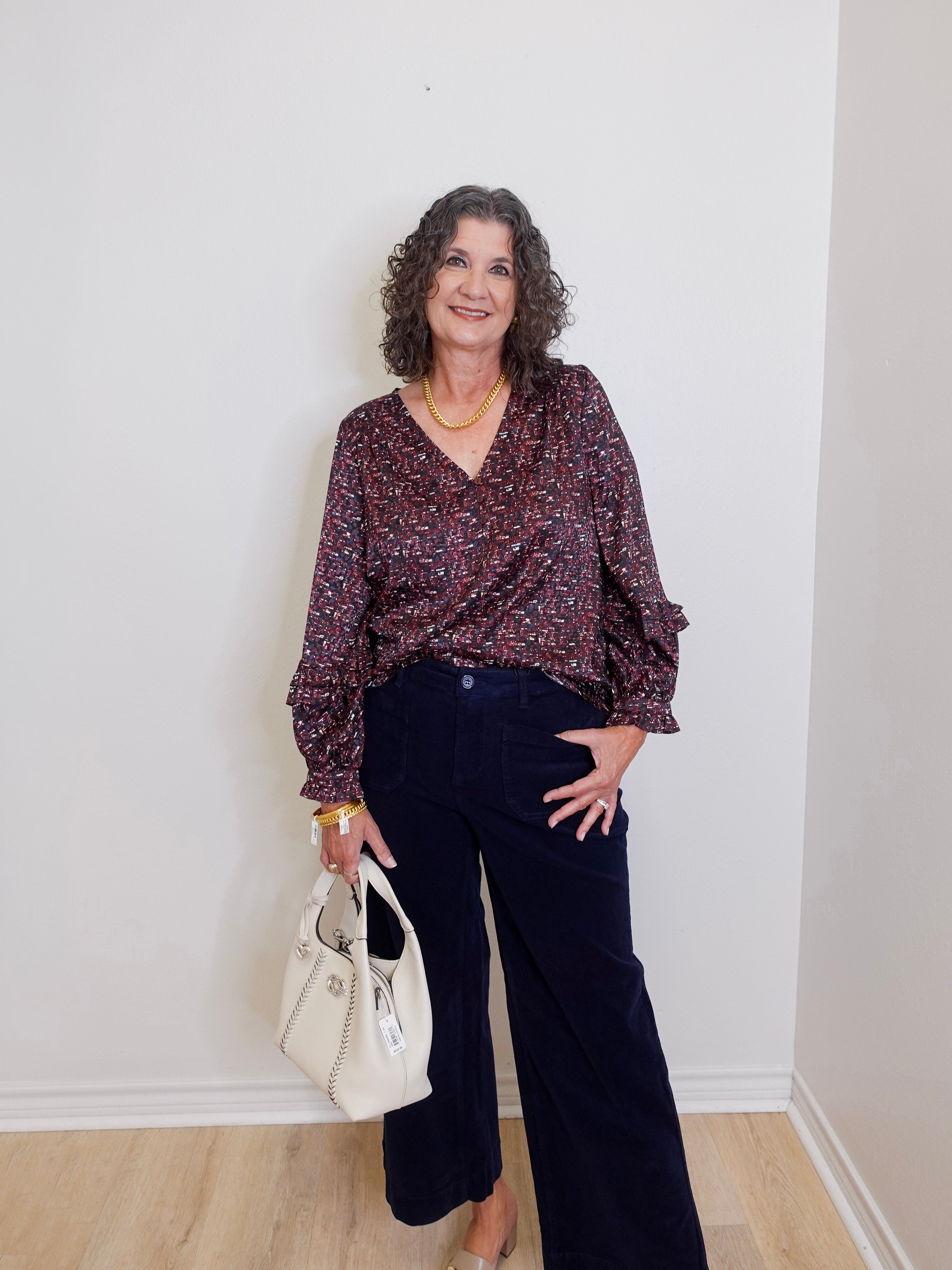 Woman wearing a patterned blouse and dark pants, holding a white handbag against a plain background