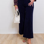 Person wearing a patterned blouse, navy pants, and gold sandals, holding a white handbag against a white wall.