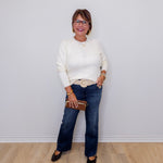 Woman wearing a white sweater and blue jeans standing against a plain white wall.