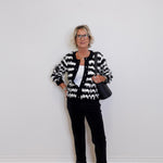 Woman wearing a black and white patterned cardigan with black pants against a white wall.