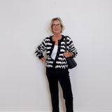 Woman wearing a black and white patterned cardigan with black pants against a white wall.