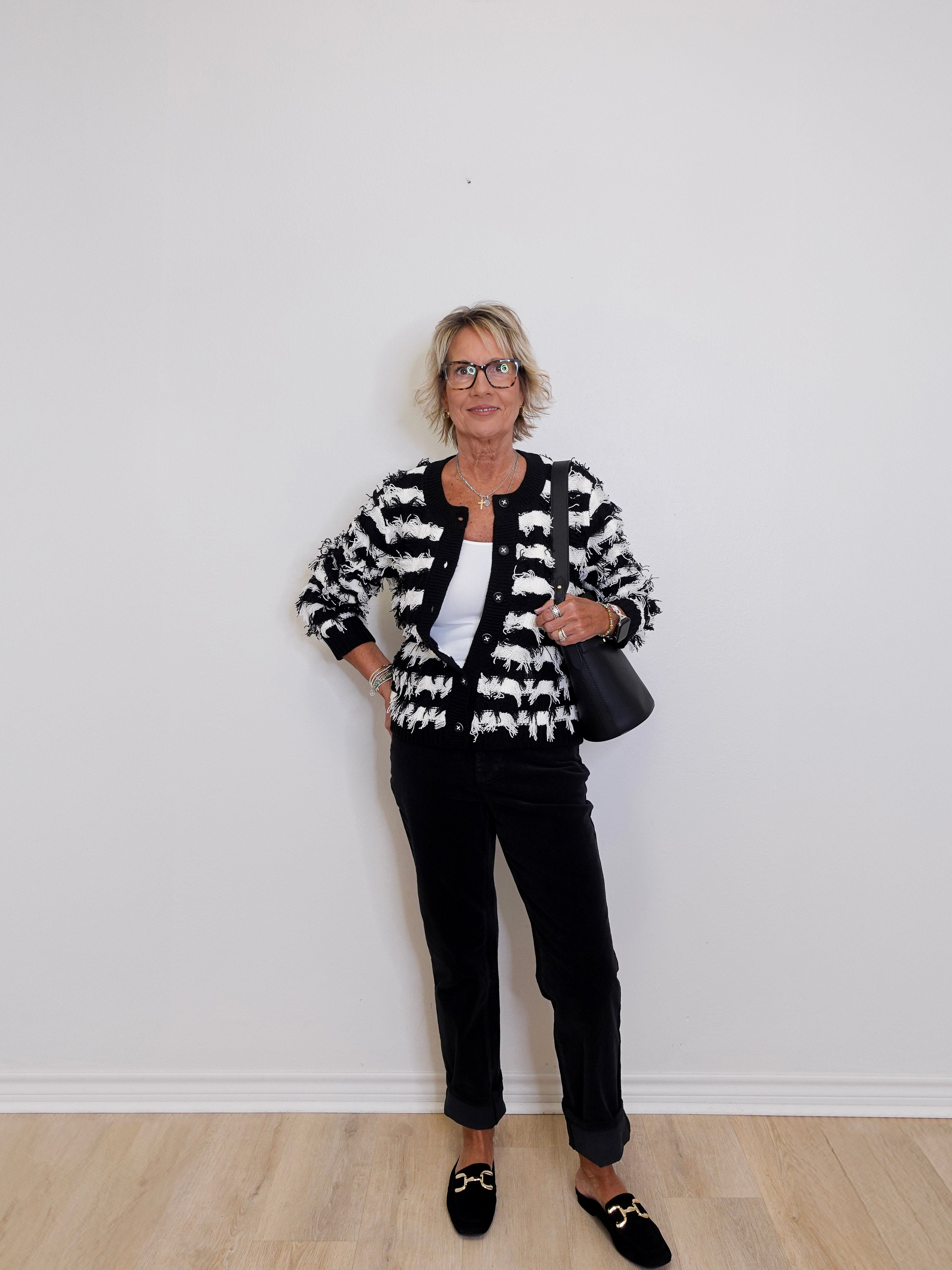 Woman wearing a black and white patterned cardigan with black pants against a white wall.
