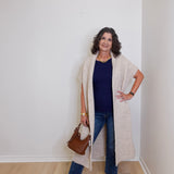 Woman wearing a long beige cardigan, blue jeans, and white sneakers in a room with a white wall.