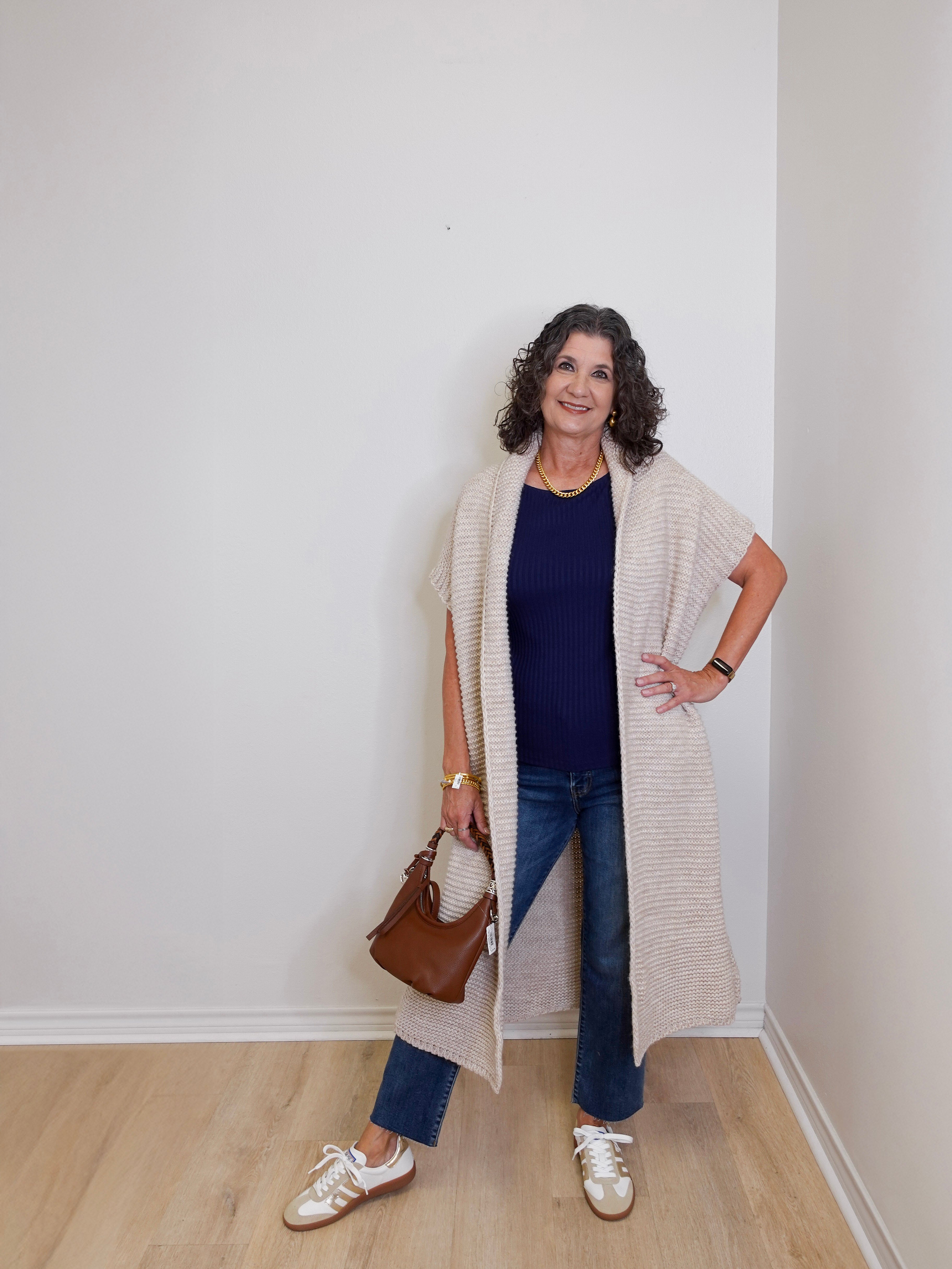 Woman wearing a long beige cardigan, blue jeans, and white sneakers in a room with a white wall.