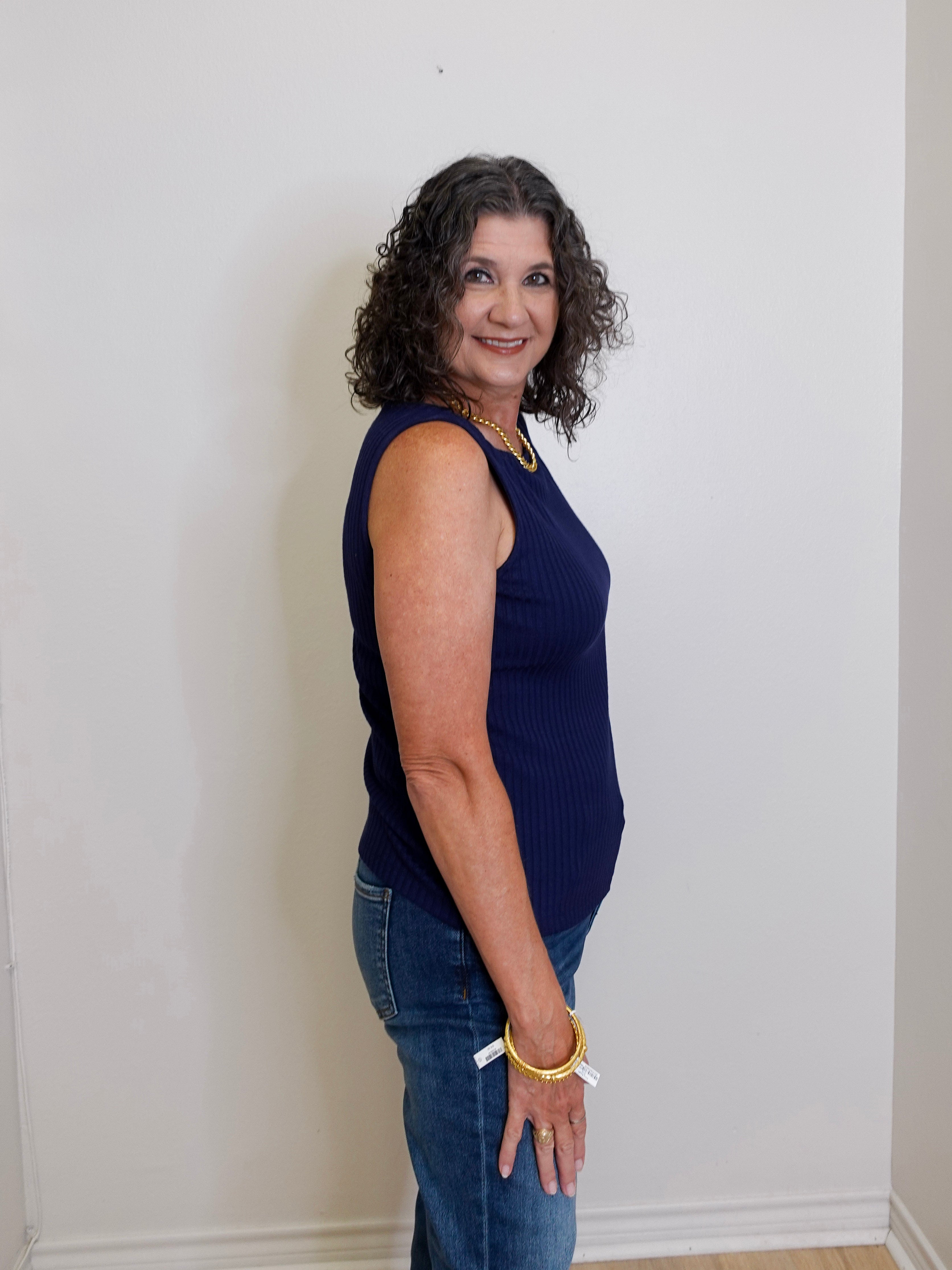 Woman wearing a navy blue sleeveless top and jeans standing against a white wall.