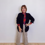 Person wearing a black jacket with colorful cuffs and trim, beige pants, and sneakers against a white wall.