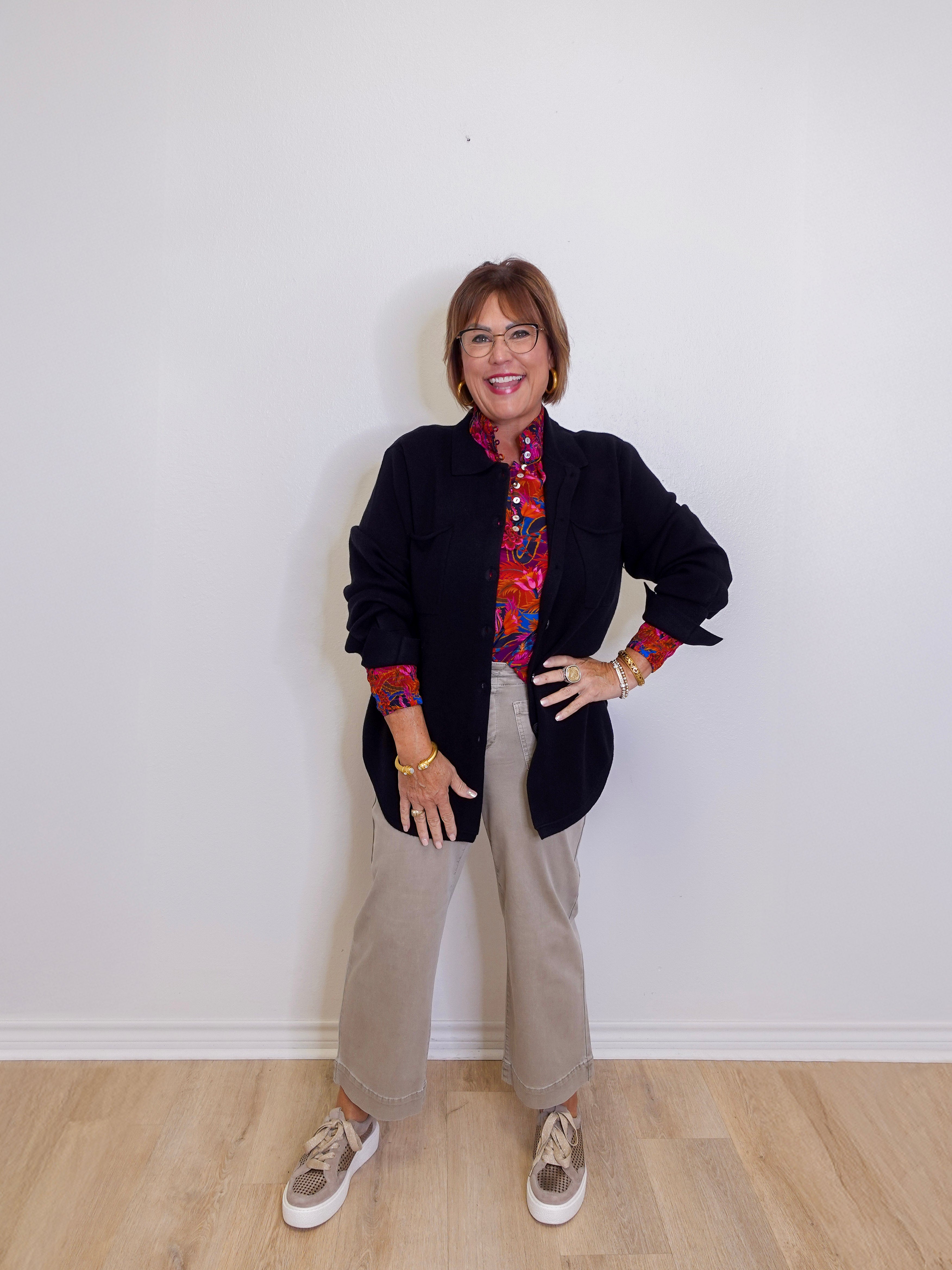 Person wearing a black jacket with colorful cuffs and trim, beige pants, and sneakers against a white wall.