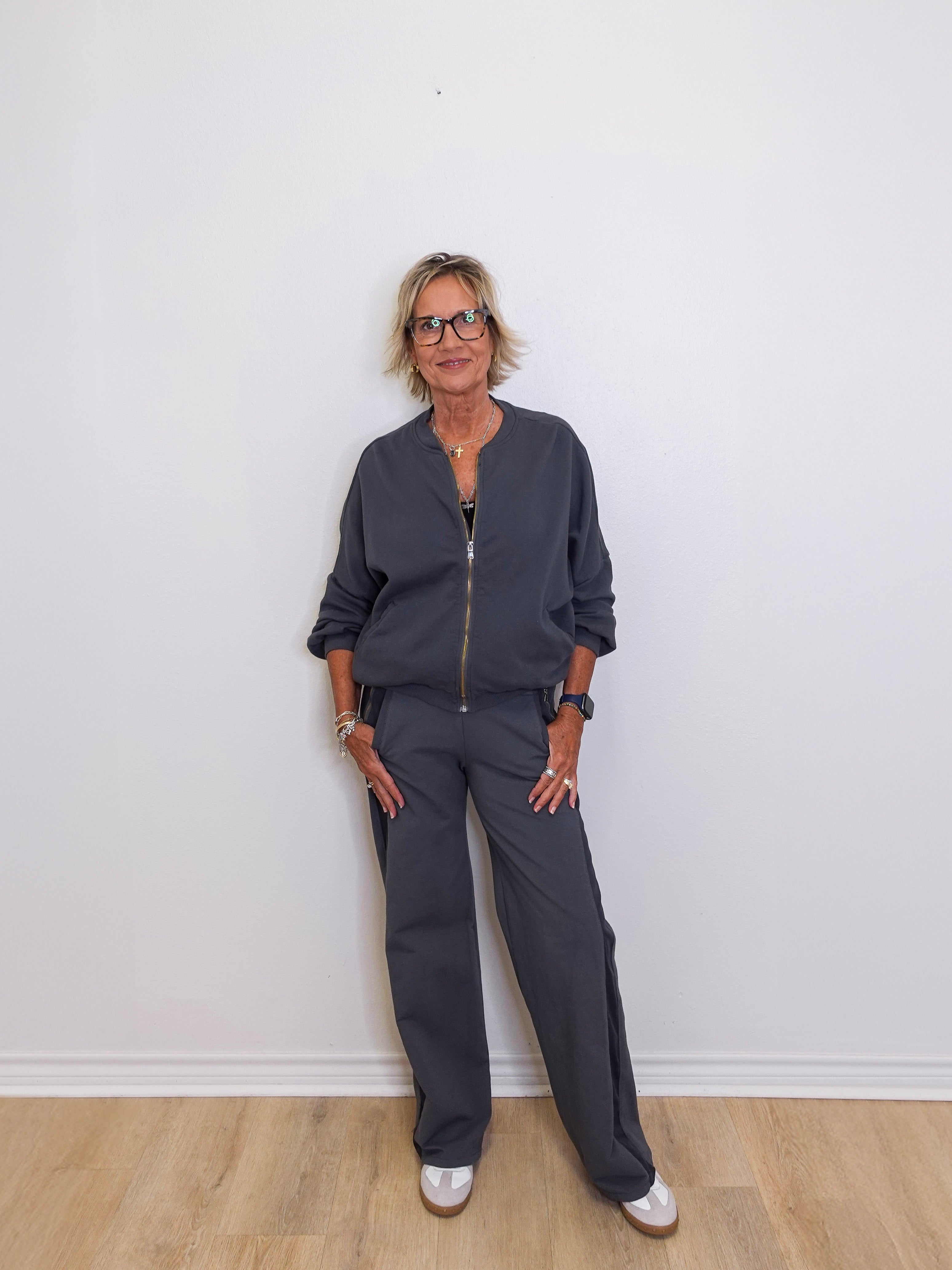 Woman wearing a dark gray outfit standing against a white wall.