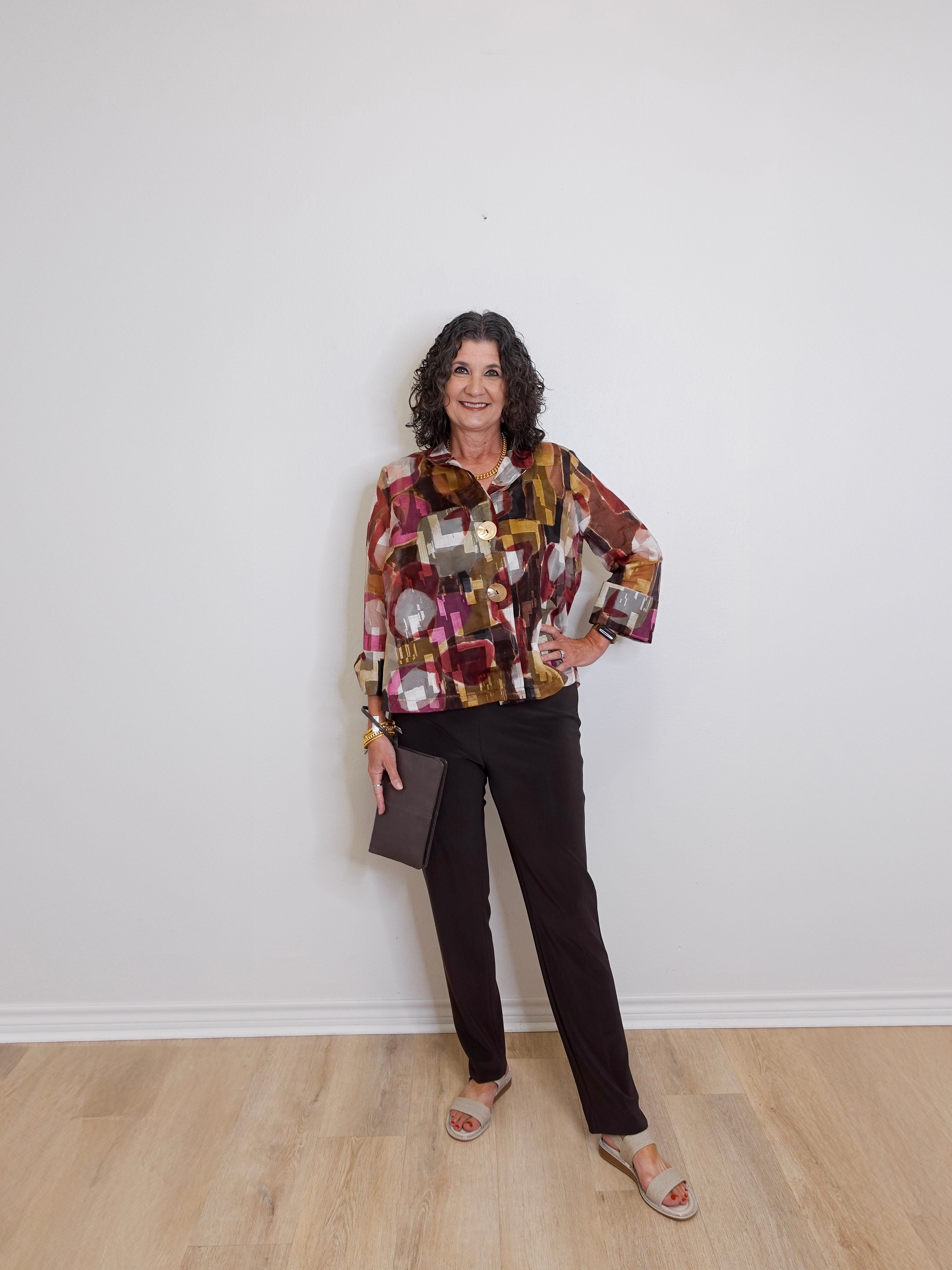 Woman wearing a patterned shirt and black pants against a white wall.
