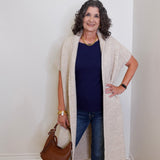 Woman wearing a long beige cardigan, blue top, and jeans, holding a brown handbag against a white wall.