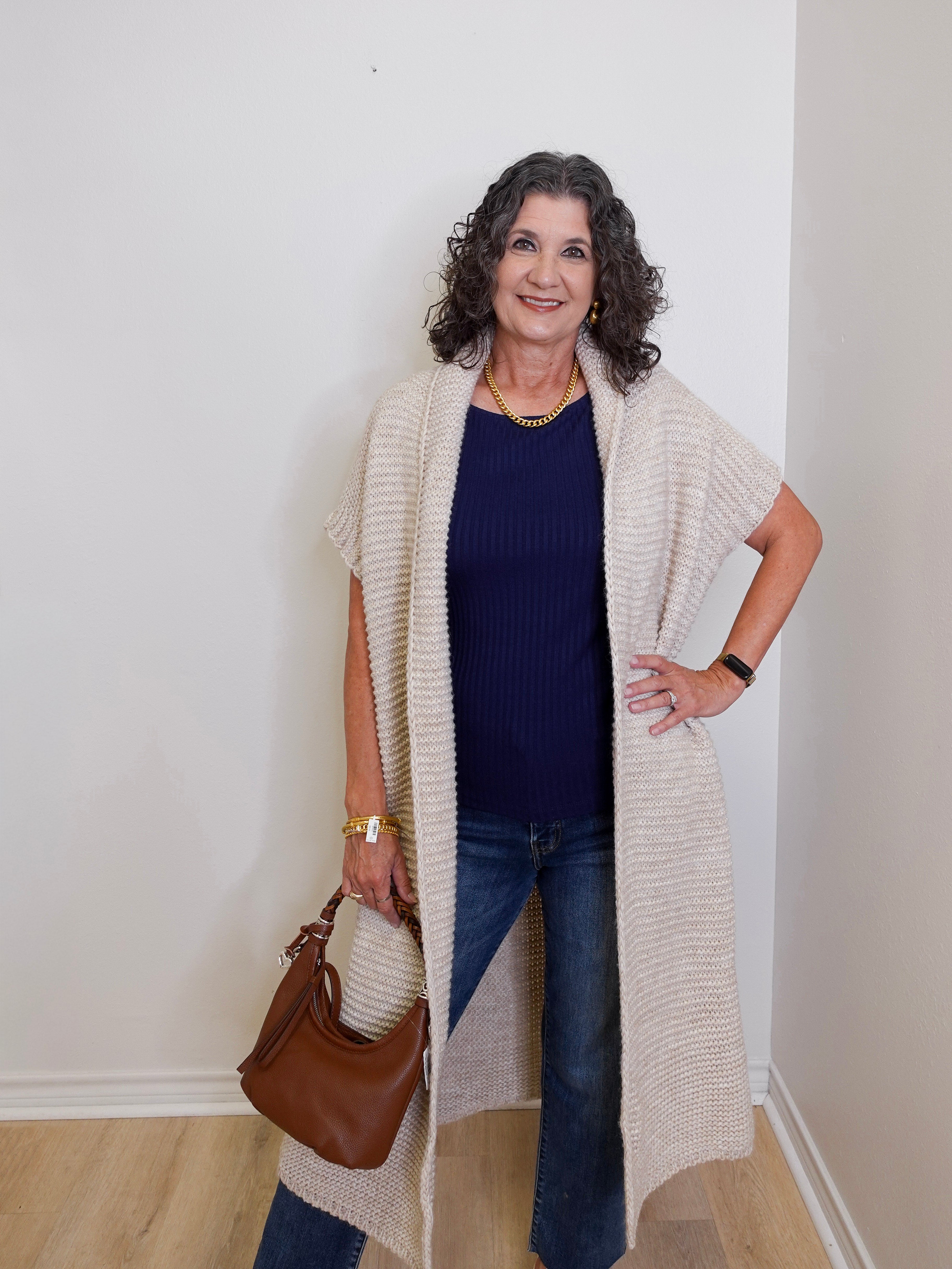Woman wearing a long beige cardigan, blue top, and jeans, holding a brown handbag against a white wall.