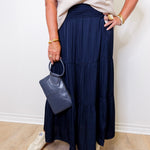 Person wearing a navy blue skirt and white top, holding a gray handbag against a white wall.