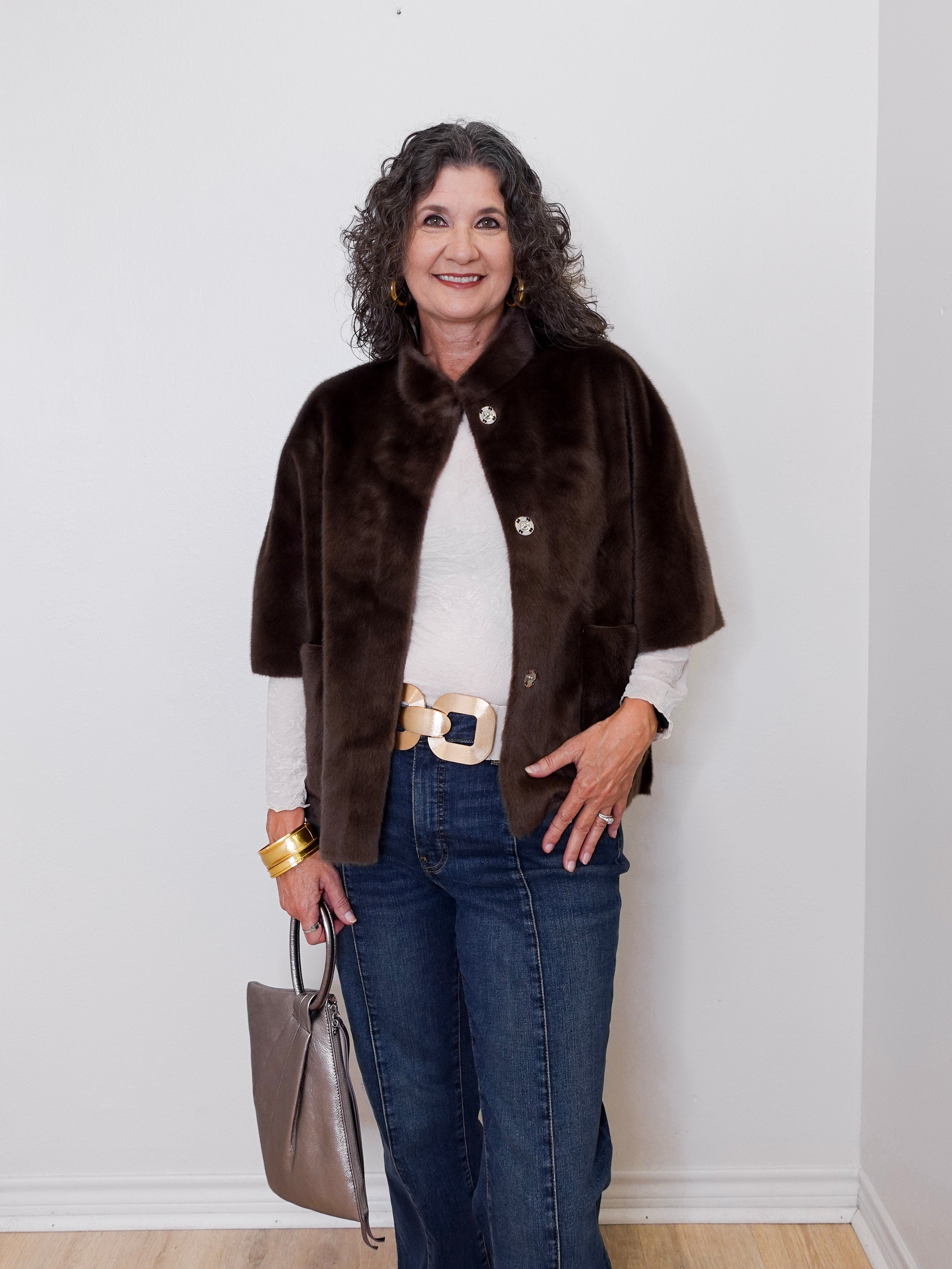 Woman wearing a brown fur coat, white shirt, blue jeans, and gold belt against a white wall.