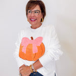 Woman wearing a white sweatshirt with a pumpkin and pink bow design, standing against a plain background.