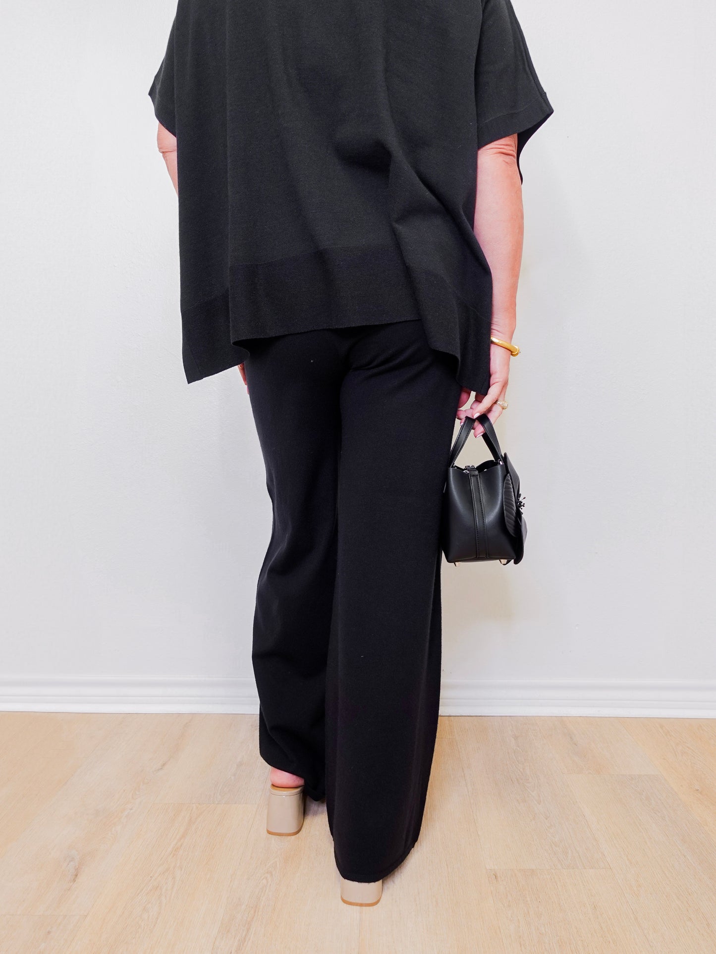 Person wearing a black outfit with wide-leg pants and holding a small black handbag against a white background.