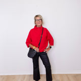 Woman in a red sweater and black pants standing against a white wall.