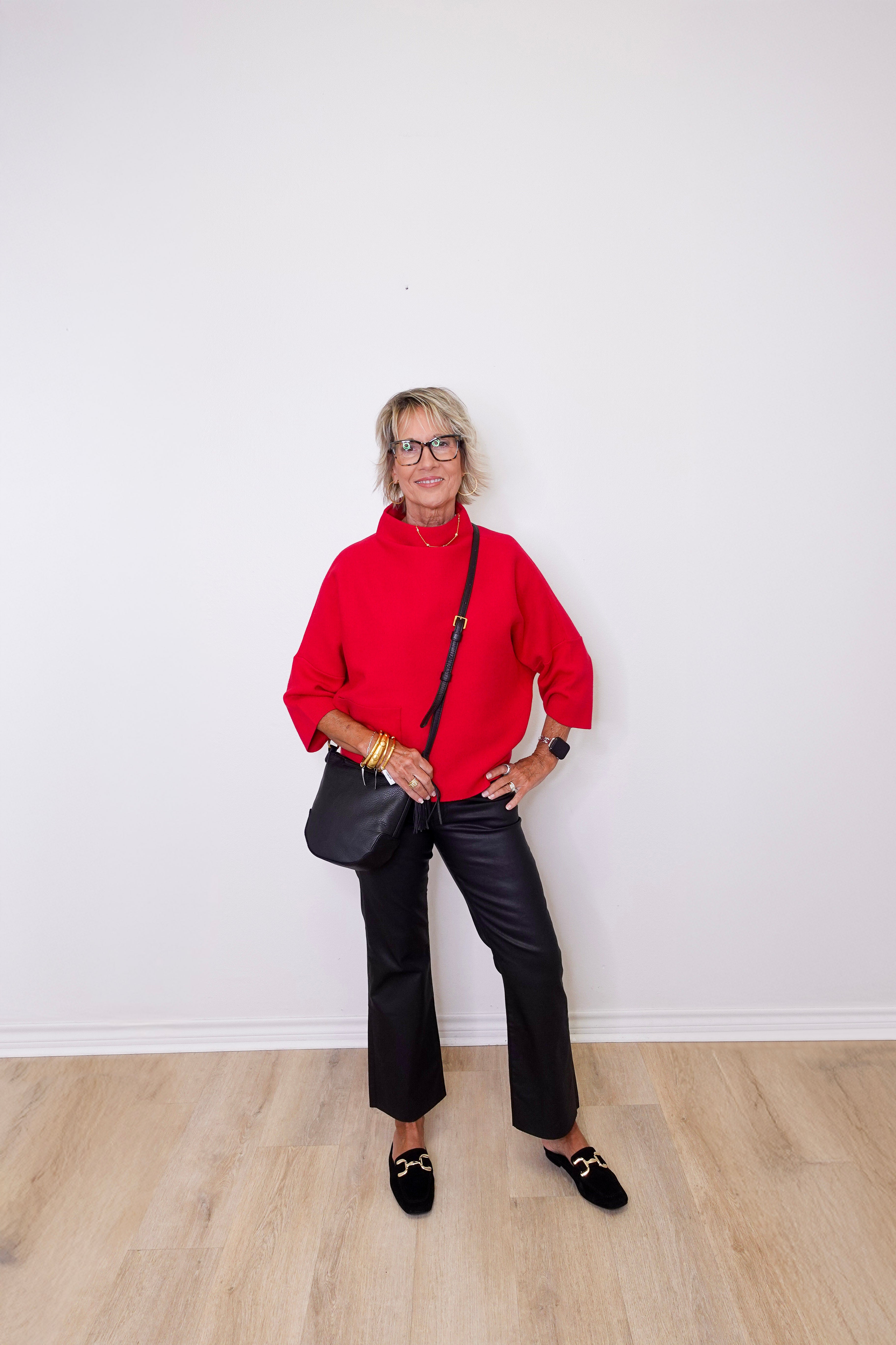 Woman in a red sweater and black pants standing against a white wall.