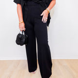 Person wearing a black outfit with wide-leg pants holding a black handbag against a white background