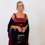 Woman wearing a burgundy dress with a black shawl and holding a black handbag against a white background